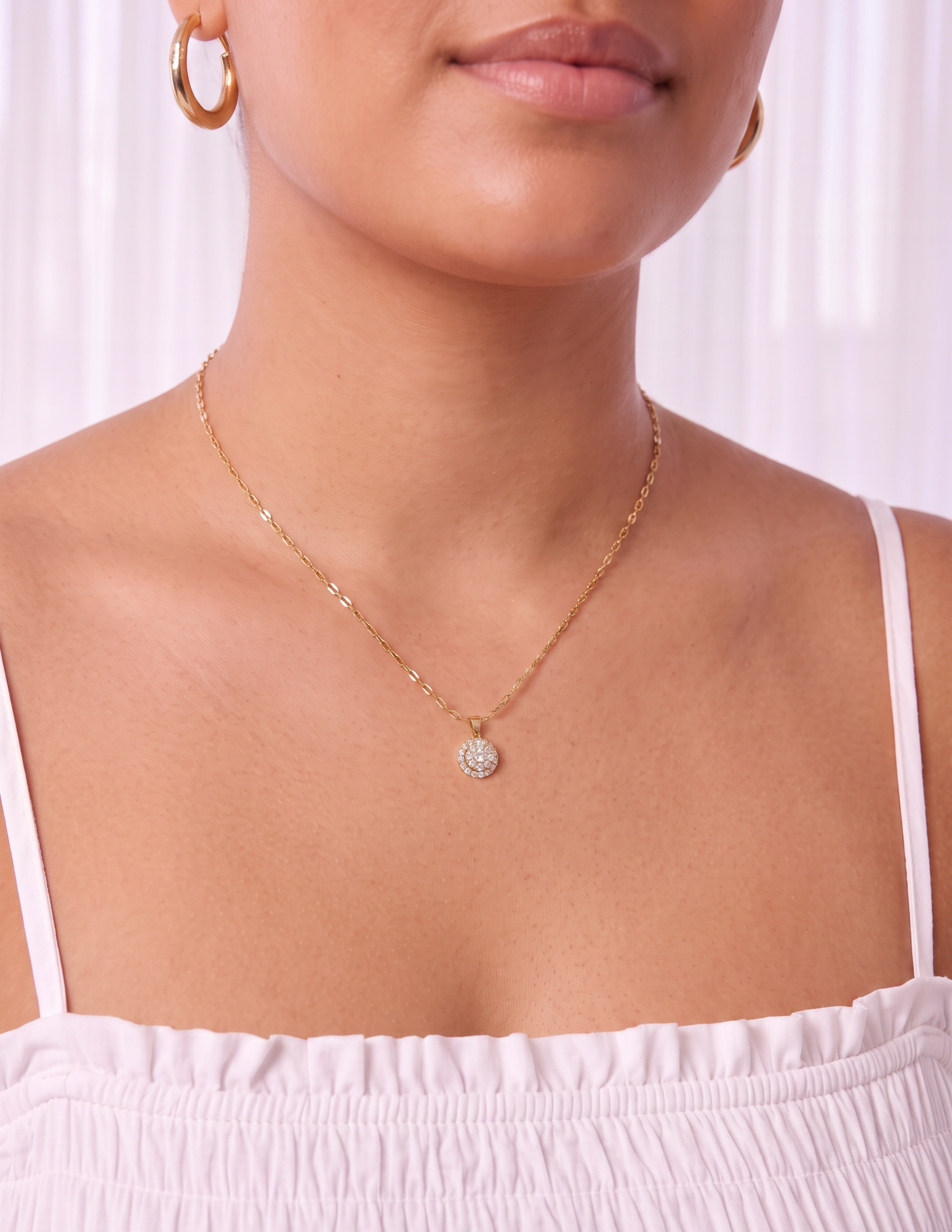 A close-up of a model wearing a delicate gold chain necklace with a small, round, clear gemstone pendant. The model is wearing a light pink off-the-shoulder top and matching gold hoop earrings, set against a soft, light-colored curtain background.