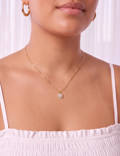 A close-up of a model wearing a delicate gold chain necklace with a small, round, clear gemstone pendant. The model is wearing a light pink off-the-shoulder top and matching gold hoop earrings, set against a soft, light-colored curtain background.