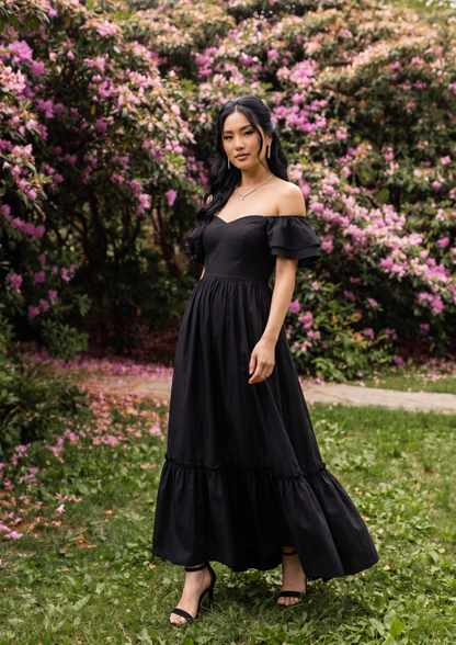 A woman wearing a black off-the-shoulder maxi dress with tiered ruffles, standing outdoors on a grassy lawn in front of a lush background of green bushes and pink flowers.