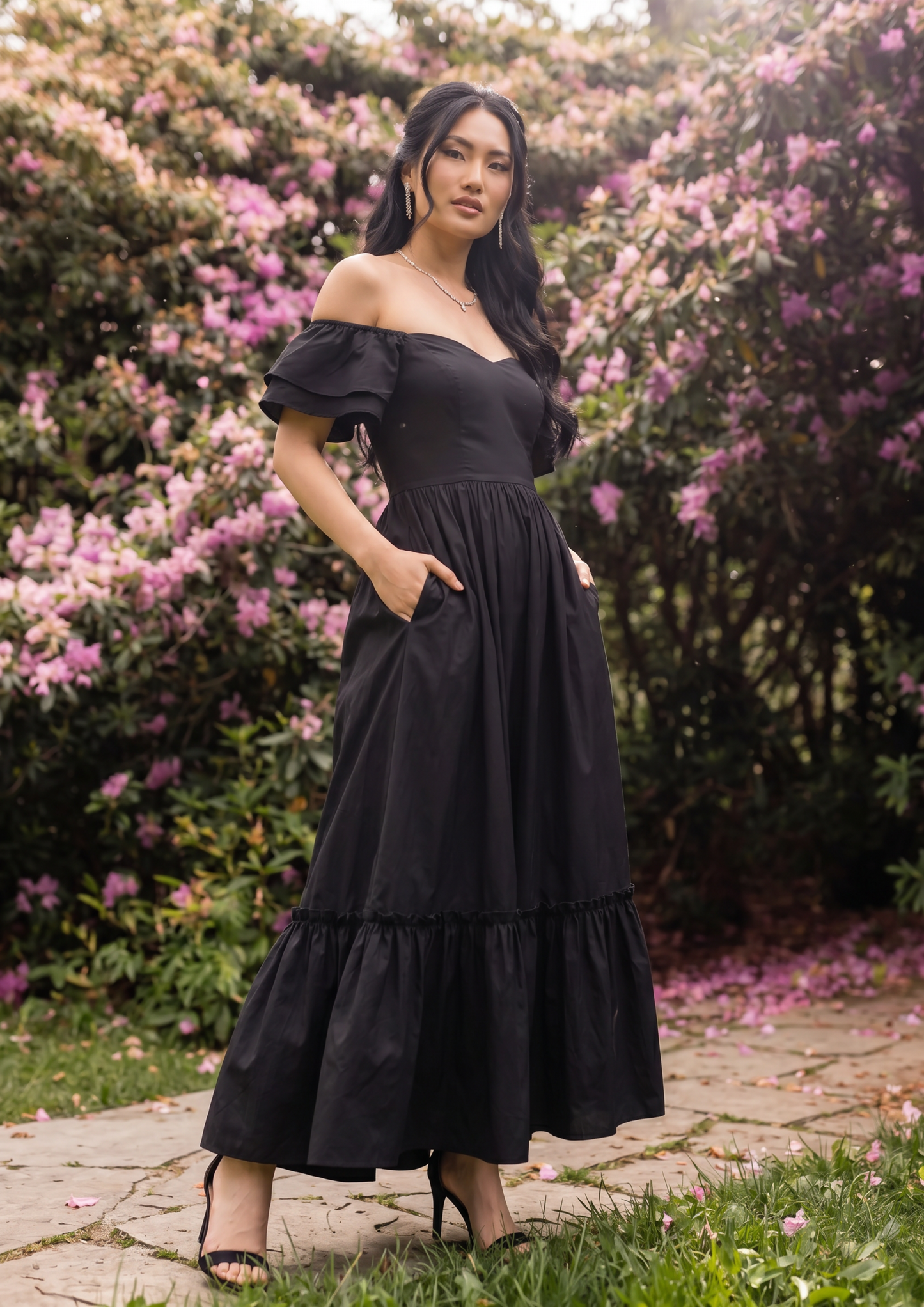 A woman wearing a black off-the-shoulder maxi dress with ruffled sleeves and tiered hem, standing outdoors on a stone path surrounded by green foliage and pink flowers.