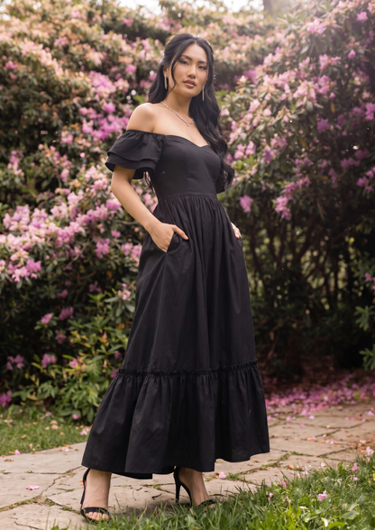 A woman wearing a black off-the-shoulder maxi dress with ruffled sleeves and tiered hem, standing outdoors on a stone path surrounded by green foliage and pink flowers.