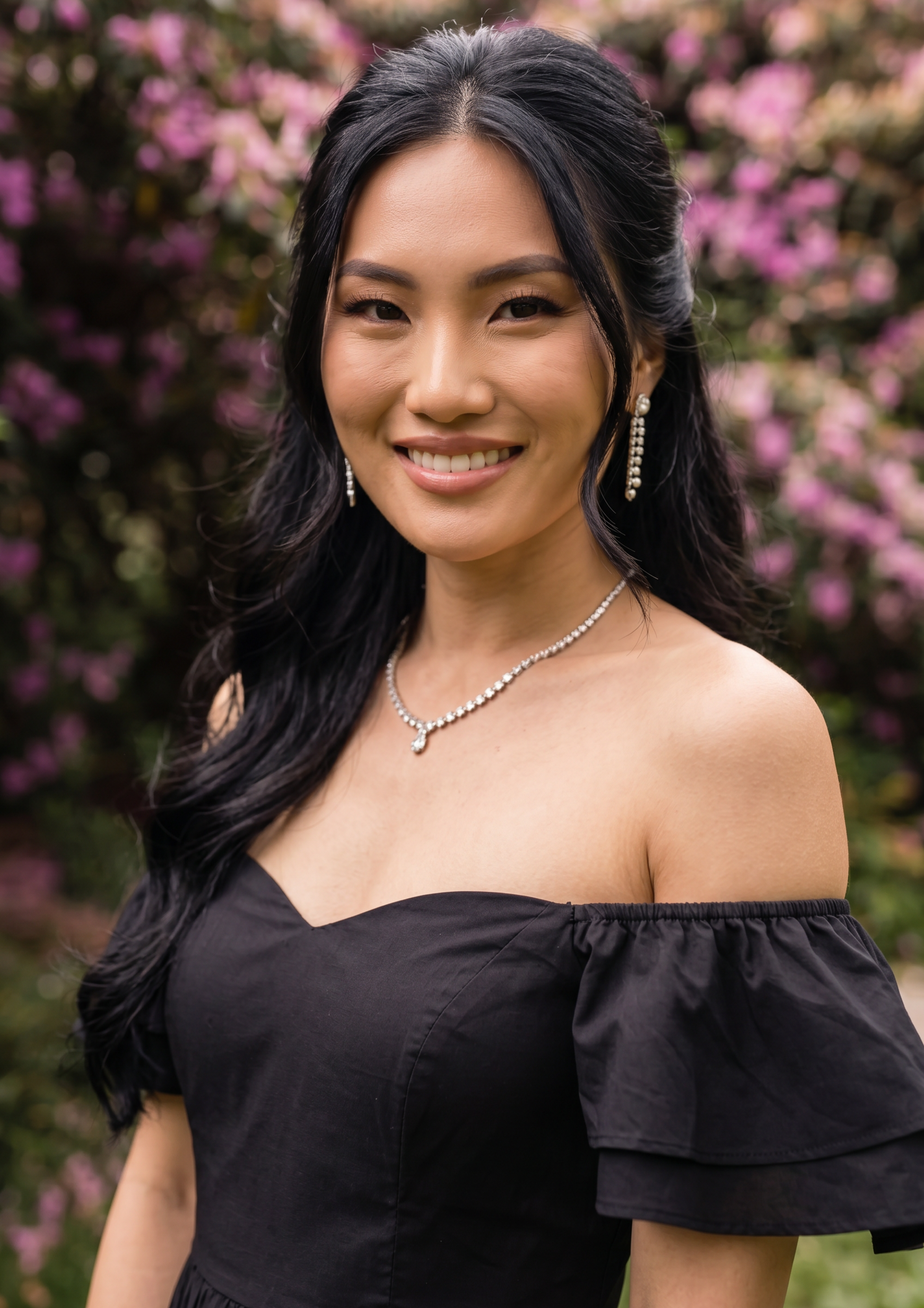 A smiling woman with long dark hair wearing a black off-the-shoulder ruffled top and silver jewelry, standing outdoors in front of a blurred background of pink flowers and green foliage.