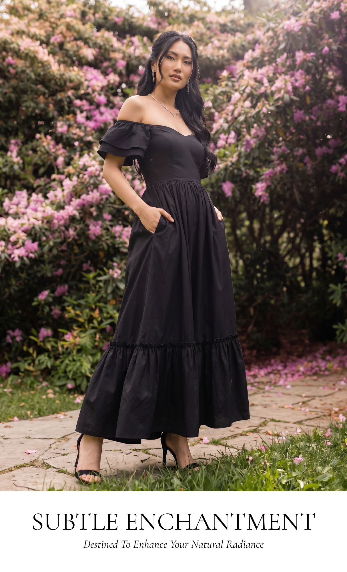 A woman wearing a black off-the-shoulder maxi dress with ruffled sleeves and tiered hem, standing outdoors on a stone path surrounded by green foliage and pink flowers. Text at the bottom reads 'SUBTLE ENCHANTMENT' with smaller copy below.