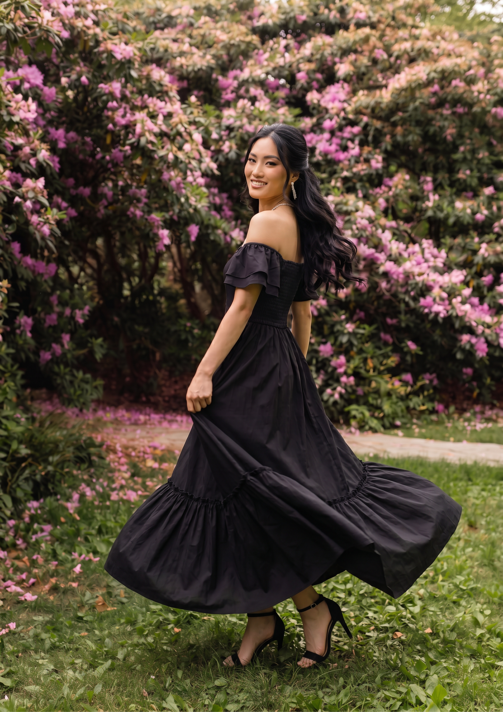 A woman wearing a black off-the-shoulder maxi dress with tiered ruffles, standing outdoors on a grassy lawn in front of a lush background of green bushes and pink flowers.