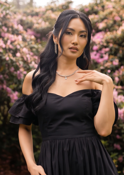 A woman with long, wavy black hair models a black off-the-shoulder dress with a sweetheart neckline in an outdoor setting with pink flowers in the background.