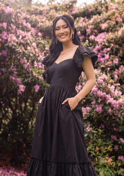 A woman wearing a light pink off-the-shoulder midi dress stands in a lush garden filled with pink, purple, and white flowers. The dress has a fitted bodice, ruffled sleeves, and a tiered skirt.