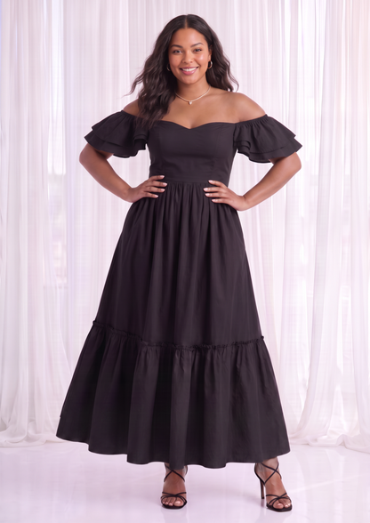 A woman modeling a black off-the-shoulder maxi dress with a sweetheart neckline, puffed short sleeves, a fitted waist, and a tiered skirt, standing against a white curtain backdrop.