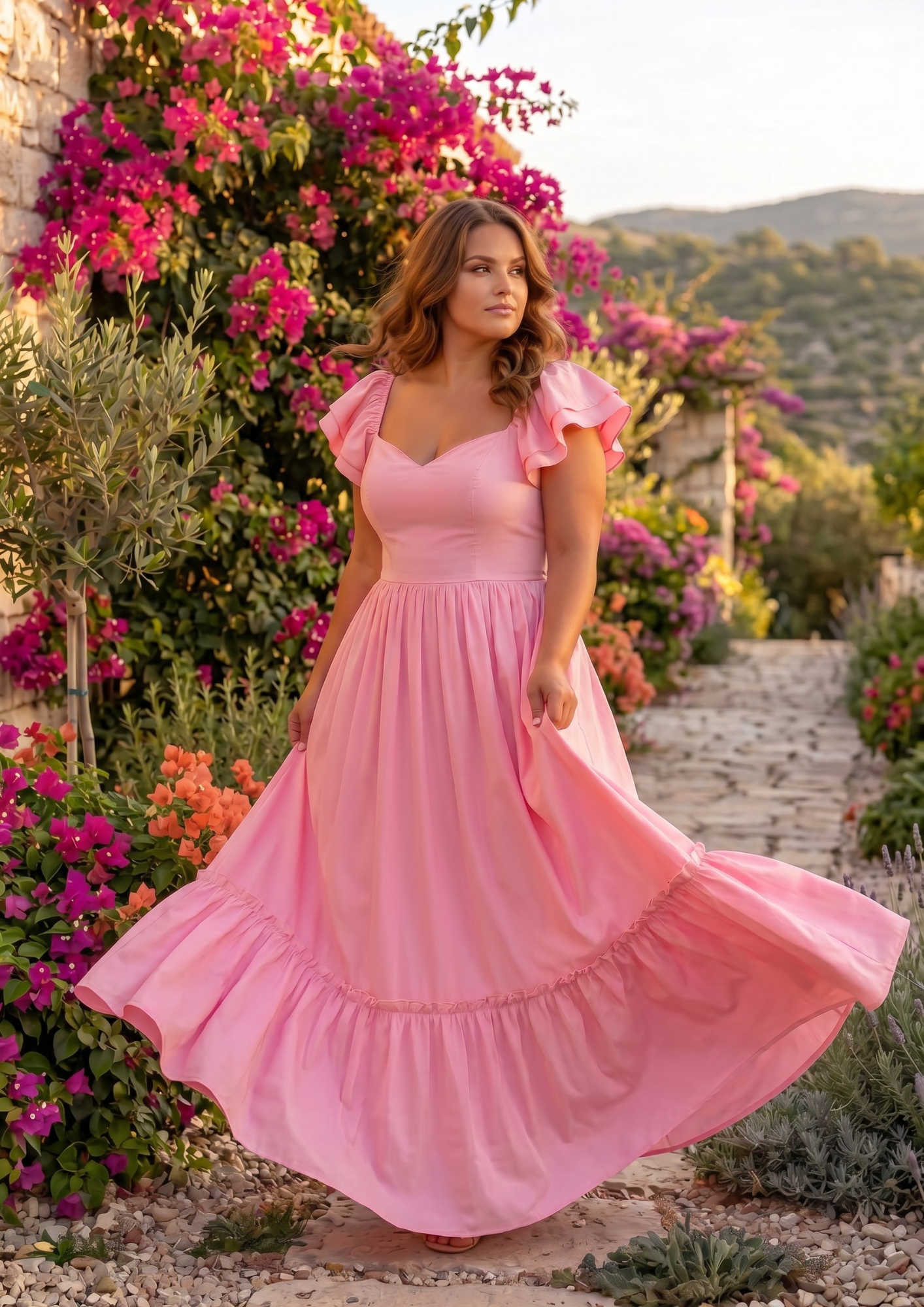 A woman wearing a flowing pink maxi dress stands on a stone path surrounded by lush greenery and bright pink flowers. The dress has ruffled short sleeves, a V-neckline, and a tiered skirt that flares out as she holds it.