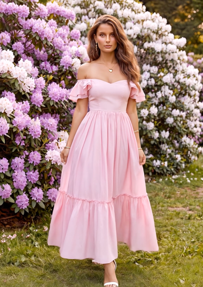 A woman wearing a light pink off-the-shoulder maxi dress with a tiered skirt, standing outdoors in front of lush greenery and pink and white flowering bushes.