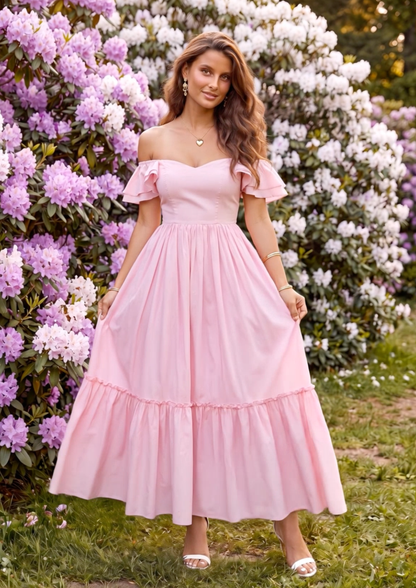 A woman wearing a light pink off-the-shoulder maxi dress with a tiered skirt, standing outdoors in front of lush green bushes with pink and white flowers.