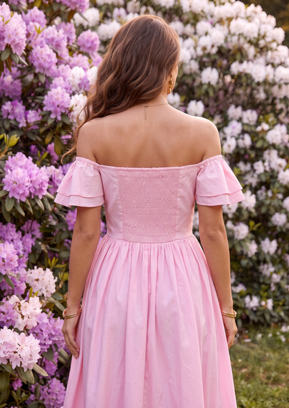 A woman viewed from behind wearing a light pink off-the-shoulder midi dress with a structured lace bodice and flared skirt, standing in front of a garden background of pink and white rhododendron flowers.