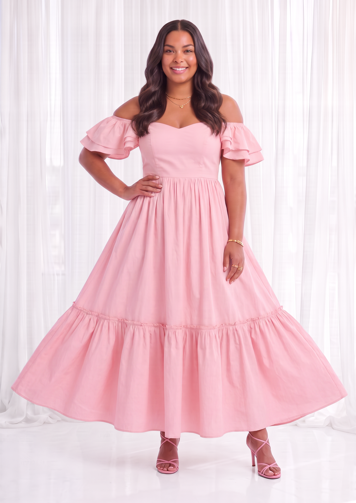 A woman modeling a light pink off-the-shoulder maxi dress with a tiered skirt, standing against a white curtain backdrop.
