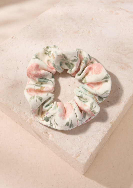 A fabric scrunchie with a soft floral print in pink, sage green, and cream, displayed on a textured beige stone block.