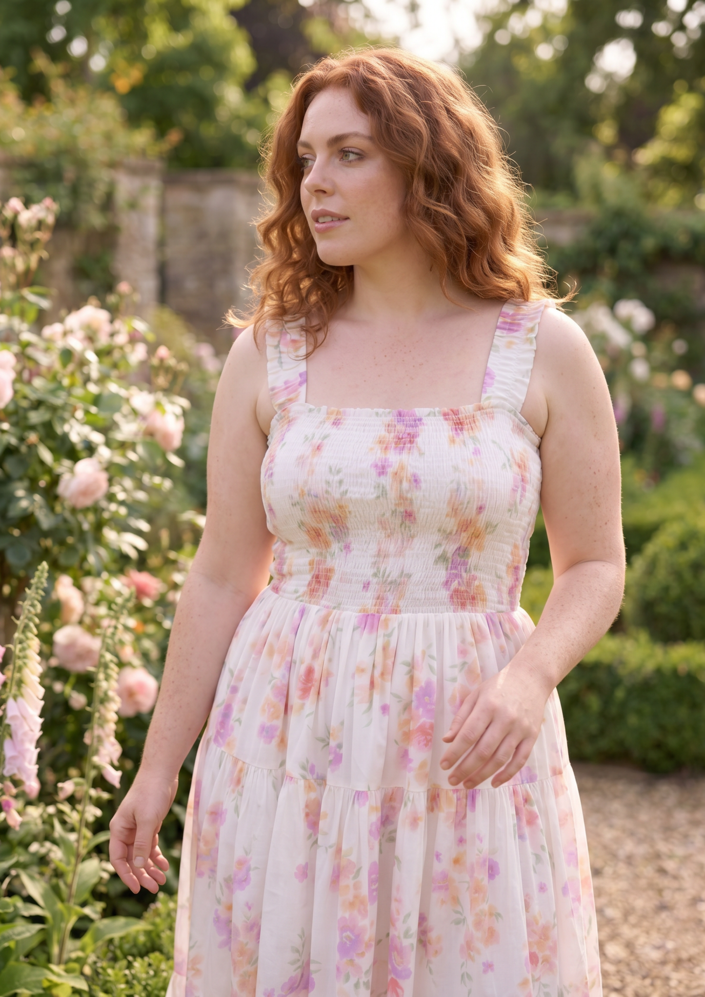 A woman with wavy red hair wearing a sleeveless tiered midi dress with a pink and yellow floral print, standing in a sunny garden with roses.