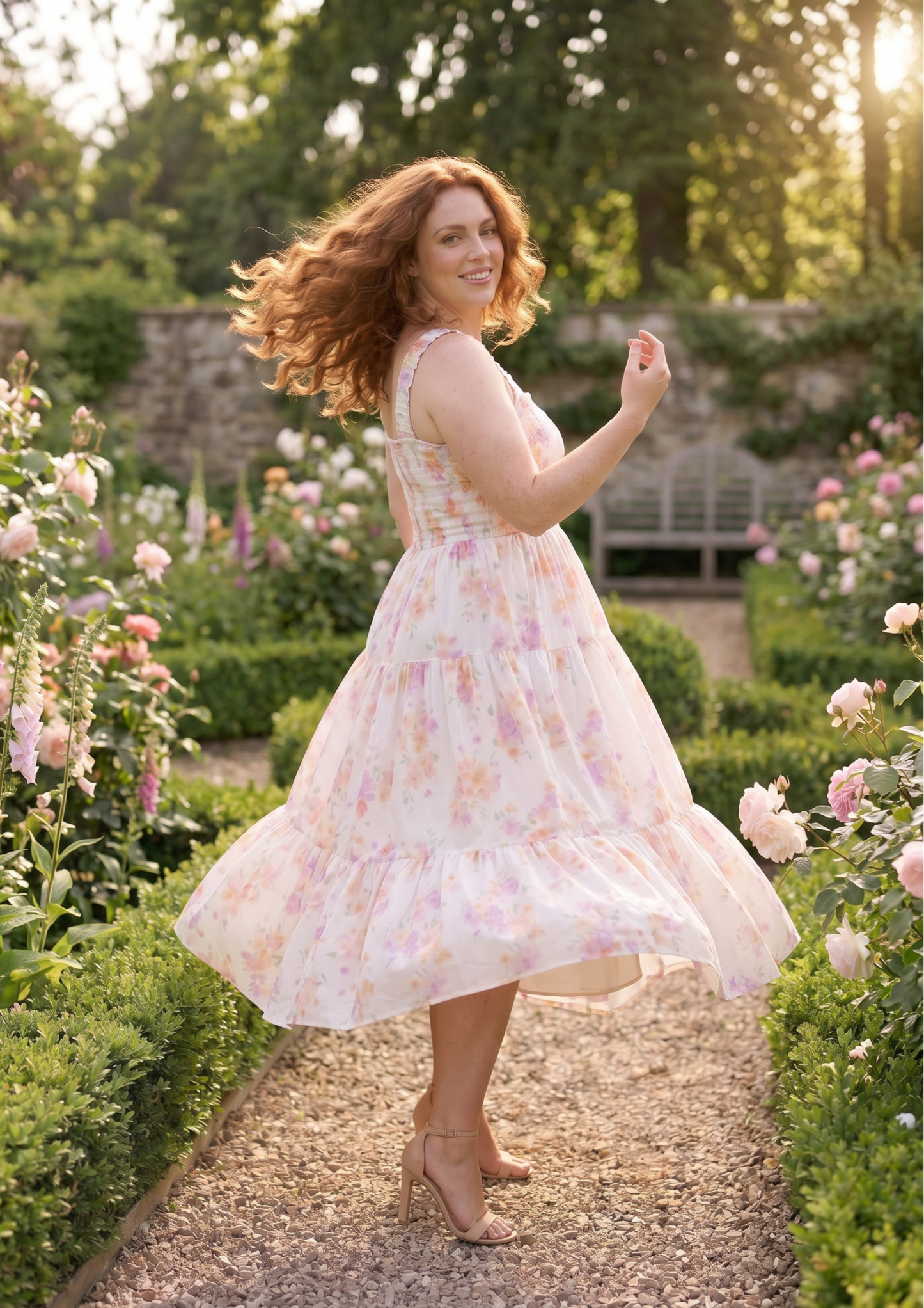 A woman with wavy red hair stands in a sunlit garden wearing a sleeveless tiered midi dress with a soft pink and peach floral print on a white base. She is captured mid-turn with her dress flowing, surrounded by blooming roses and green hedges.