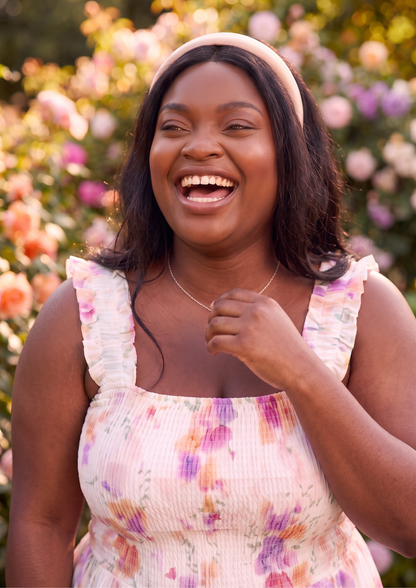 A woman smiling broadly outdoors in front of a blurred rose garden. She is wearing a light pink sleeveless top with a purple and orange floral print, a matching pink headband, and a delicate gold necklace.