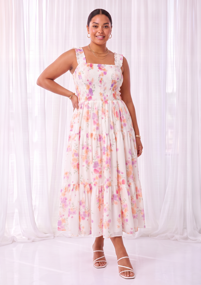 A woman modeling a sleeveless, tiered midi dress with a white base and a pastel pink, purple, and yellow floral pattern, standing in front of white sheer curtains.