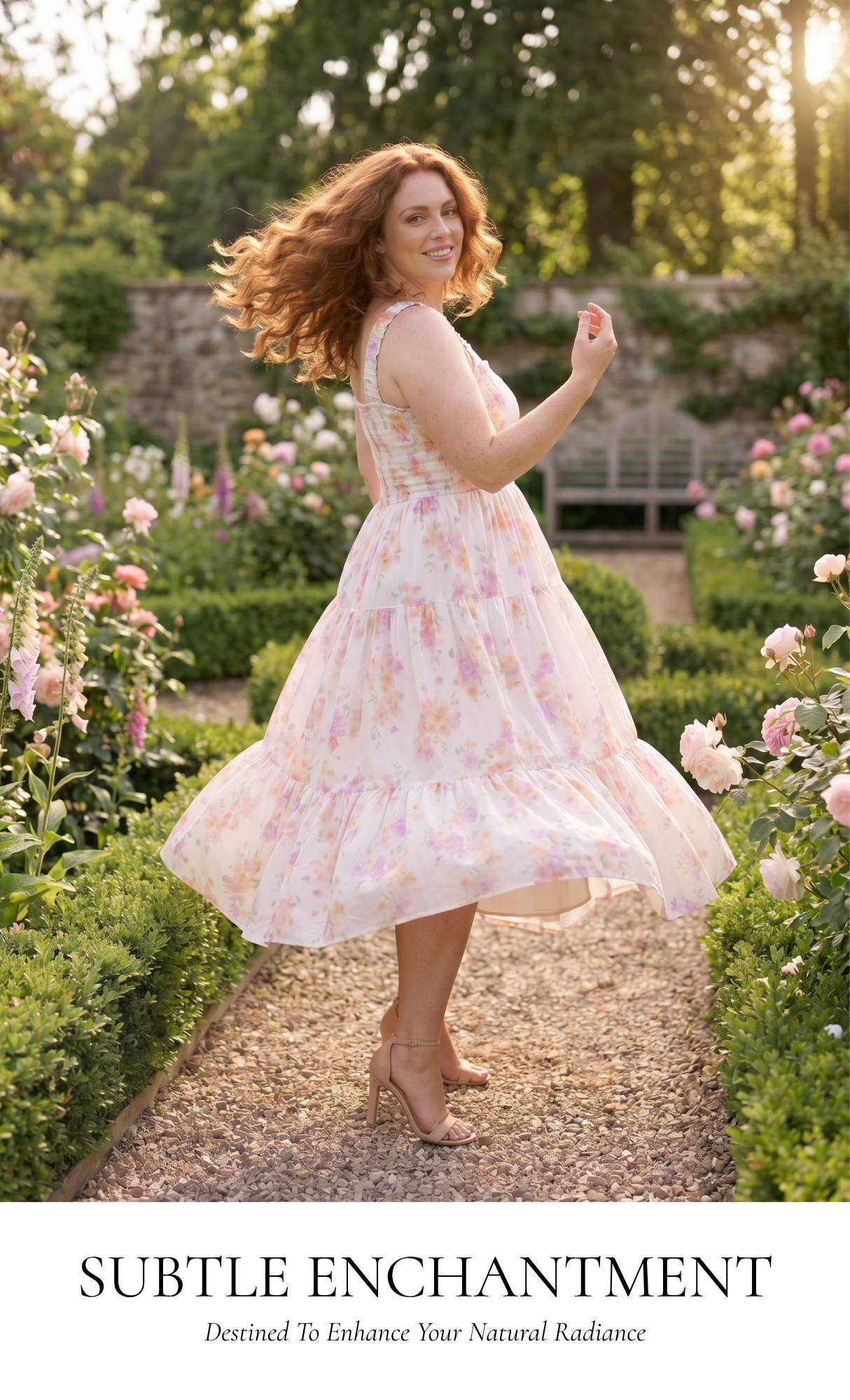 A woman with wavy red hair stands in a sunlit garden wearing a sleeveless tiered midi dress with a soft pink and peach floral print on a white base. She is captured mid-turn with her dress flowing, surrounded by blooming roses and green hedges.