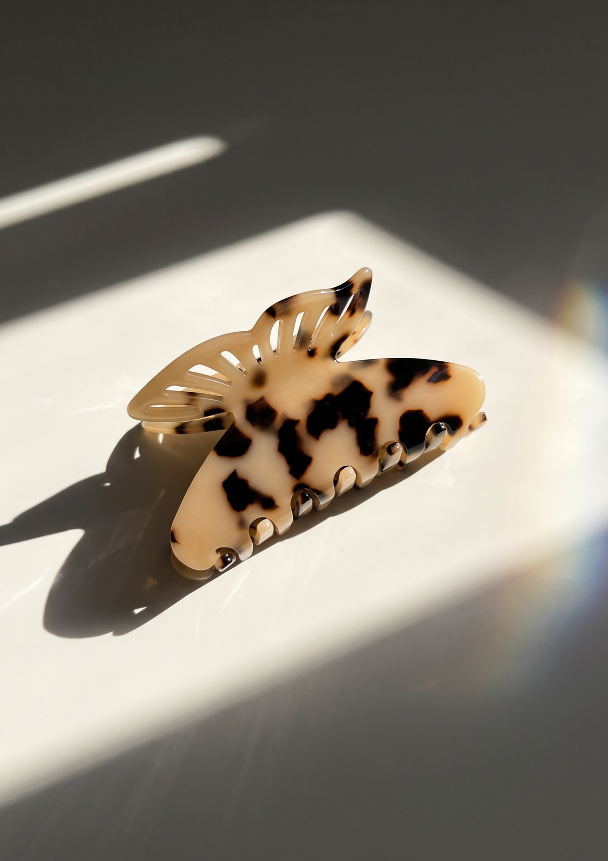 A tortoiseshell hair claw clip with cream, black, and brown mottled spots, resting on a white surface under dramatic diagonal sunlight.