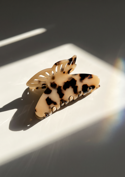 A tortoiseshell hair claw clip with cream, black, and brown mottled spots, resting on a white surface under dramatic diagonal sunlight.