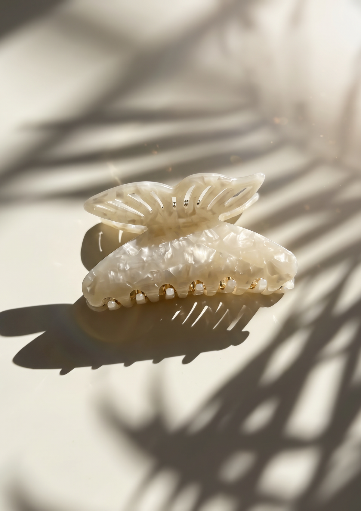 A translucent ivory hair claw clip with a textured, organic pattern, resting on a light beige surface under warm sunlight with palm leaf shadows.