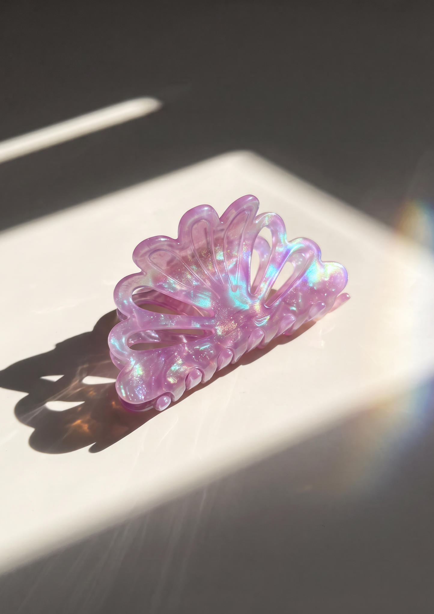 A translucent lavender purple hair claw clip with an iridescent holographic shimmer, resting on a white surface under direct sunlight.