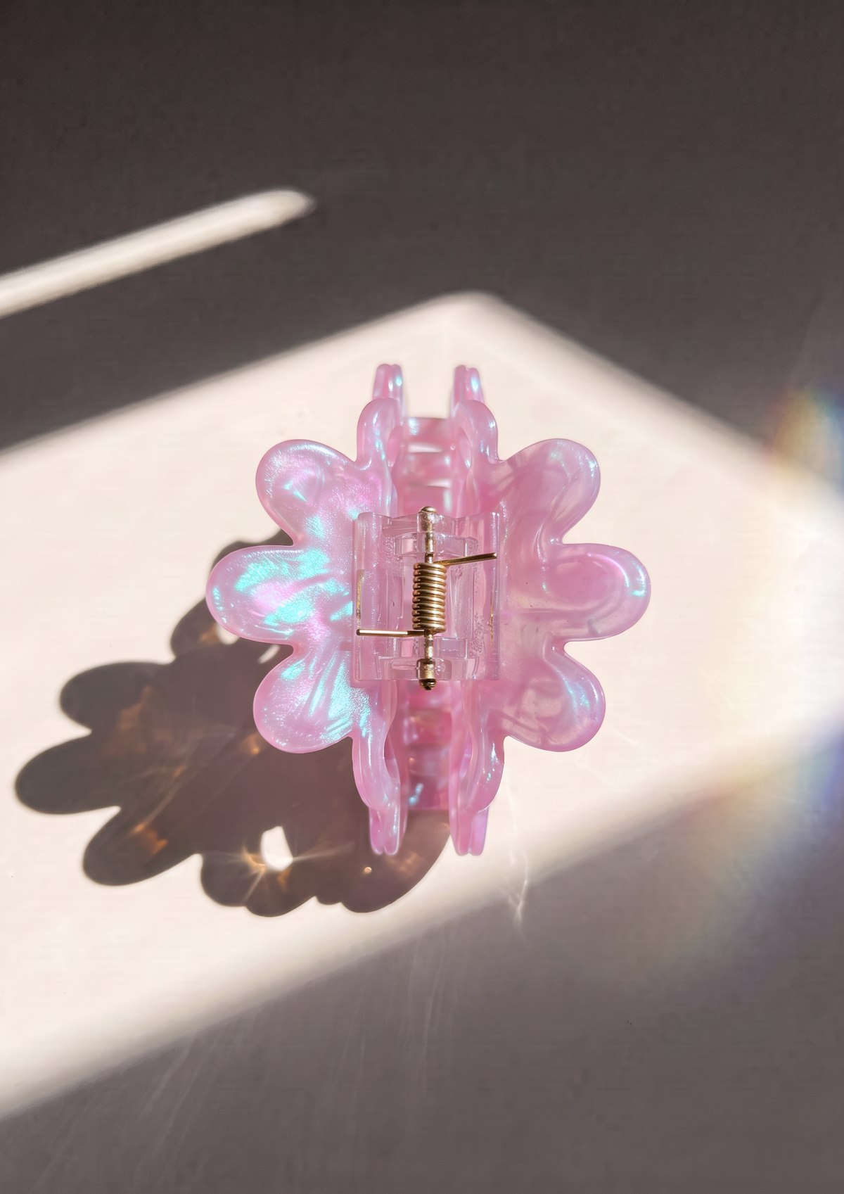 A translucent pink flower-shaped hair claw clip with an iridescent finish and a gold metal spring, resting on a white surface with dramatic sunlight and shadow.