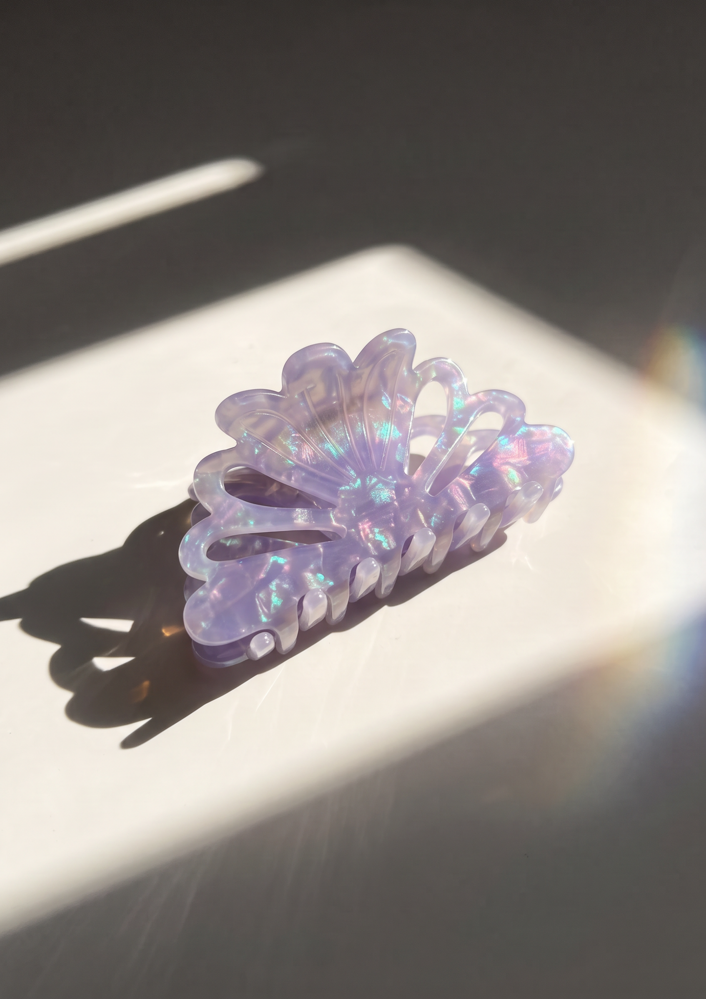 A translucent lavender hair claw clip with an iridescent holographic finish, resting on a white rectangular surface under dramatic direct sunlight and shadow.