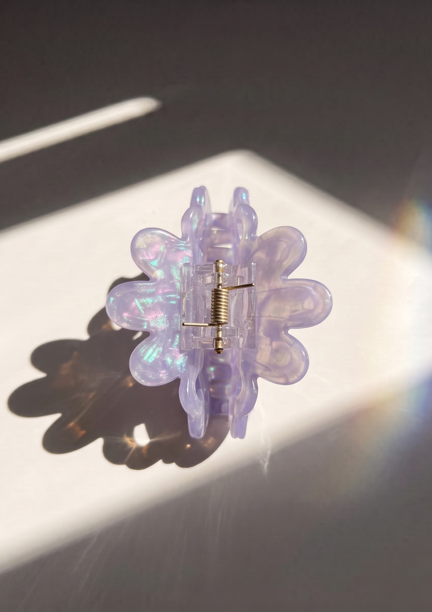A translucent lavender flower-shaped hair claw clip with an iridescent finish and a gold metal spring, photographed on a white surface with dramatic sunlight and shadow.
