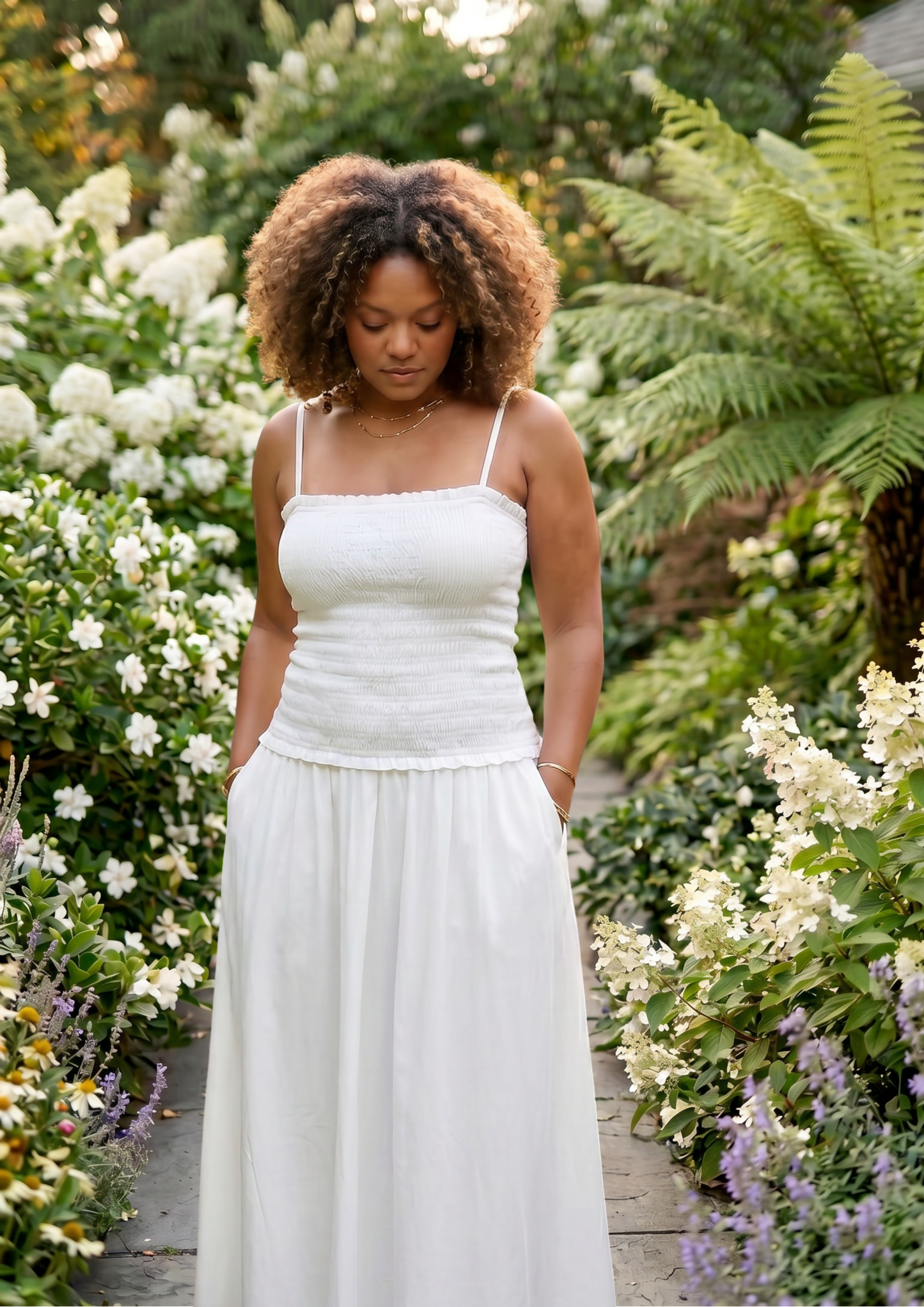 A woman with curly brown hair wearing a white spaghetti-strap maxi dress with a textured bodice and side pockets, standing in a lush garden pathway surrounded by green foliage and white flowers.