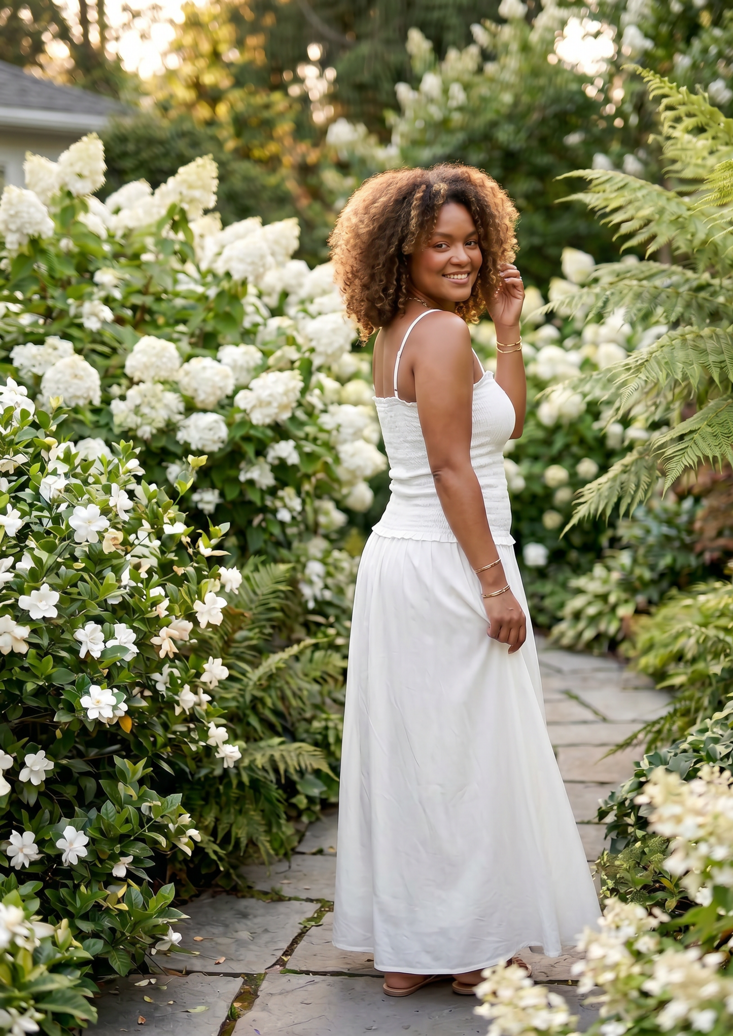 A woman wearing a white sleeveless maxi dress stands on a stone garden path surrounded by lush greenery and white flowers.