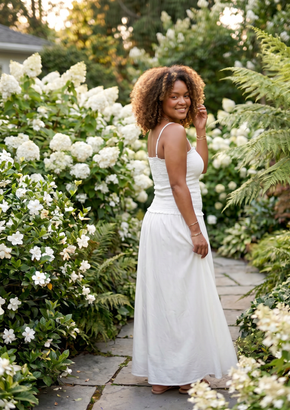 A woman wearing a white sleeveless maxi dress stands on a stone garden path surrounded by lush greenery and white flowers.