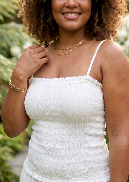 A woman wearing a white textured spaghetti-strap top, photographed outdoors with blurred green foliage in the background.