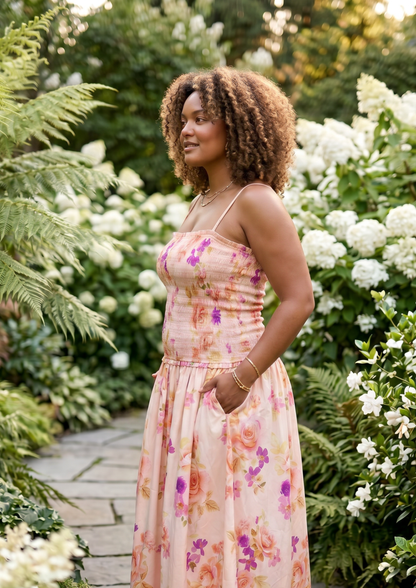 A woman with curly brown hair stands in a lush garden wearing a light peach floral maxi dress with thin spaghetti straps and a gathered waist. The background features green foliage, ferns, and white hydrangea blooms.