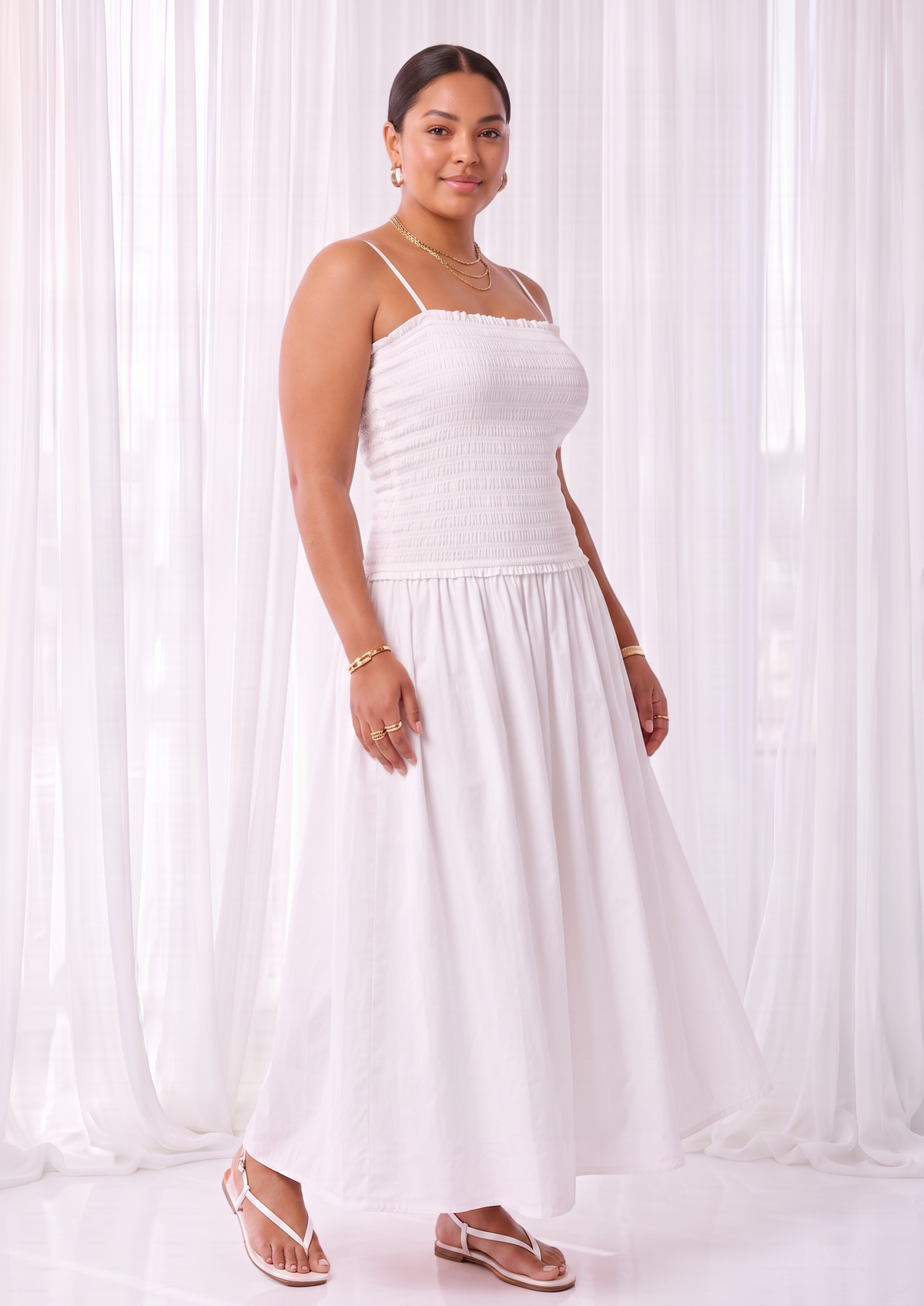 A woman modeling a white sleeveless maxi dress with thin straps and a gathered waist, standing in front of white sheer curtains.