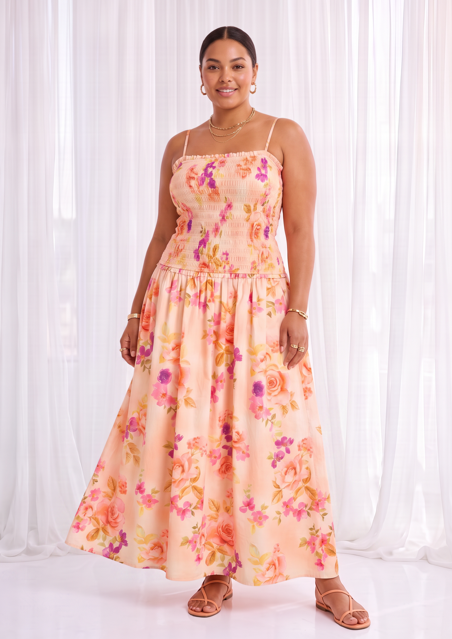 A woman modeling a peach-colored maxi dress with a vibrant pink, purple, and orange floral print, standing in front of white sheer curtains.
