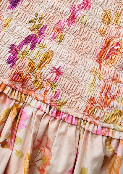Close-up of a smocked floral dress or skirt in a light blush pink fabric with a gathered ruffle and a multicolor floral print in pink, orange, yellow, and purple.