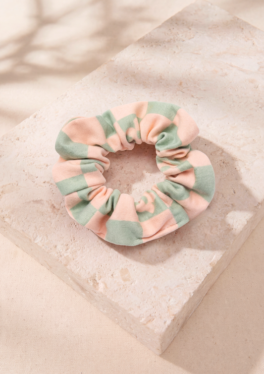 A fabric hair scrunchie with a soft pastel checkered pattern of mint green and pale pink, displayed on a textured beige stone block with soft leaf shadows.