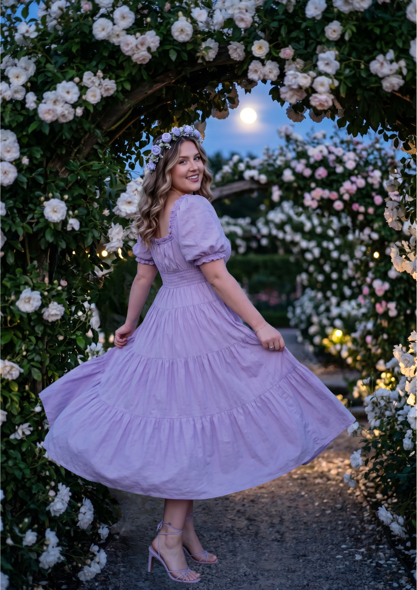 A woman wearing a lavender tiered puff-sleeve dress stands under a rose arch at dusk, holding out the skirt to show its full flow. She is wearing clear high heels and a matching flower crown.