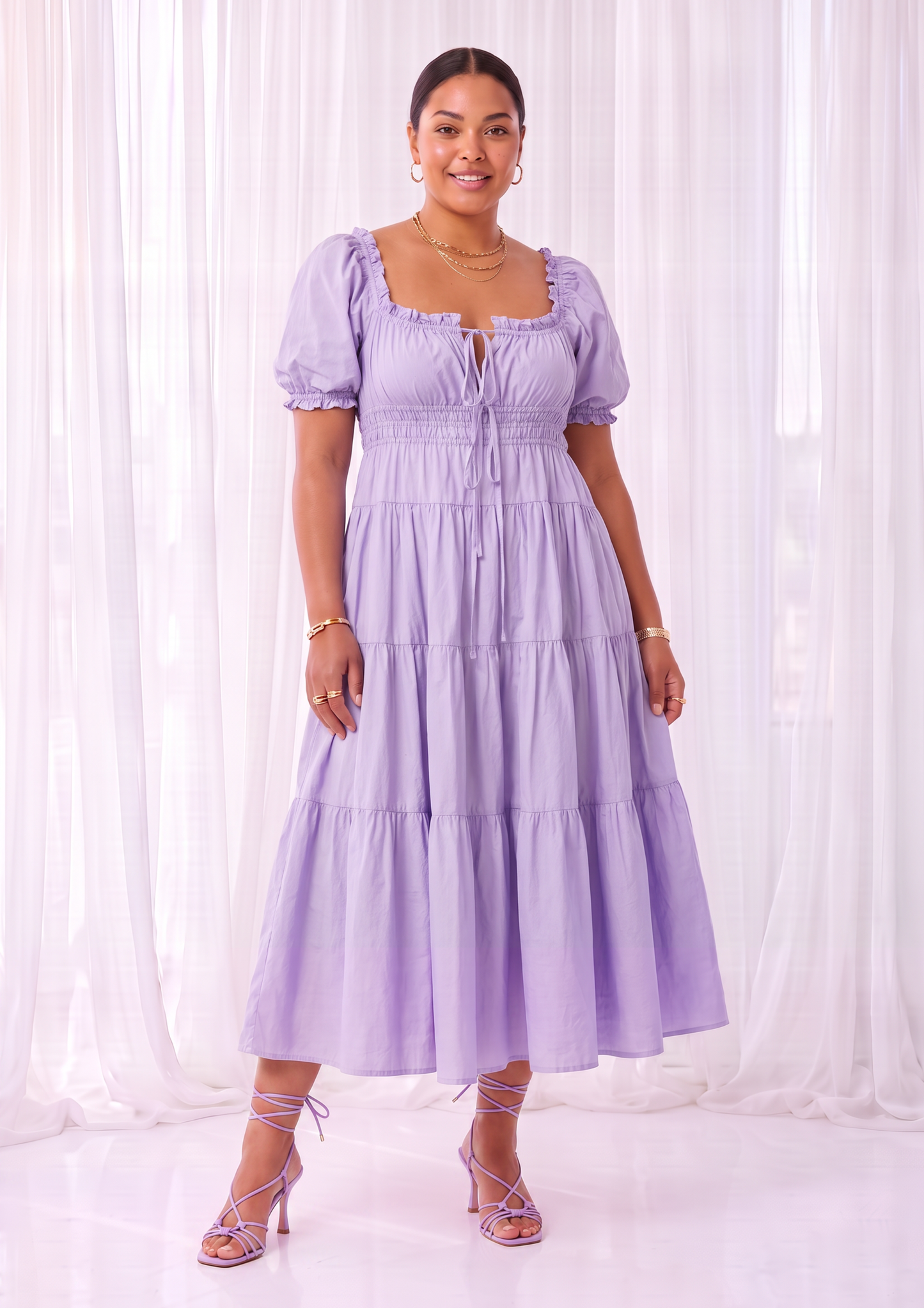 A woman modeling a light purple tiered midi dress with puff sleeves, a smocked bodice, and a front button placket, standing in front of white sheer curtains.
