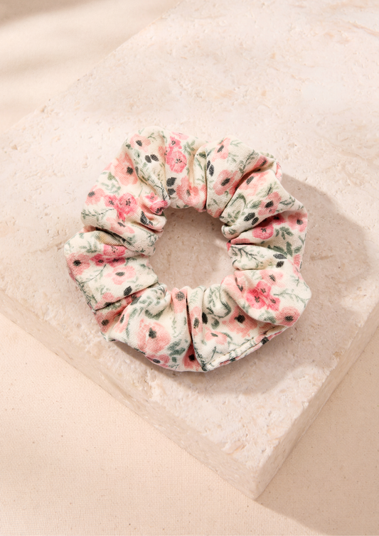 A fabric hair scrunchie with a pink and green floral pattern on a cream background, displayed on a textured beige stone block.