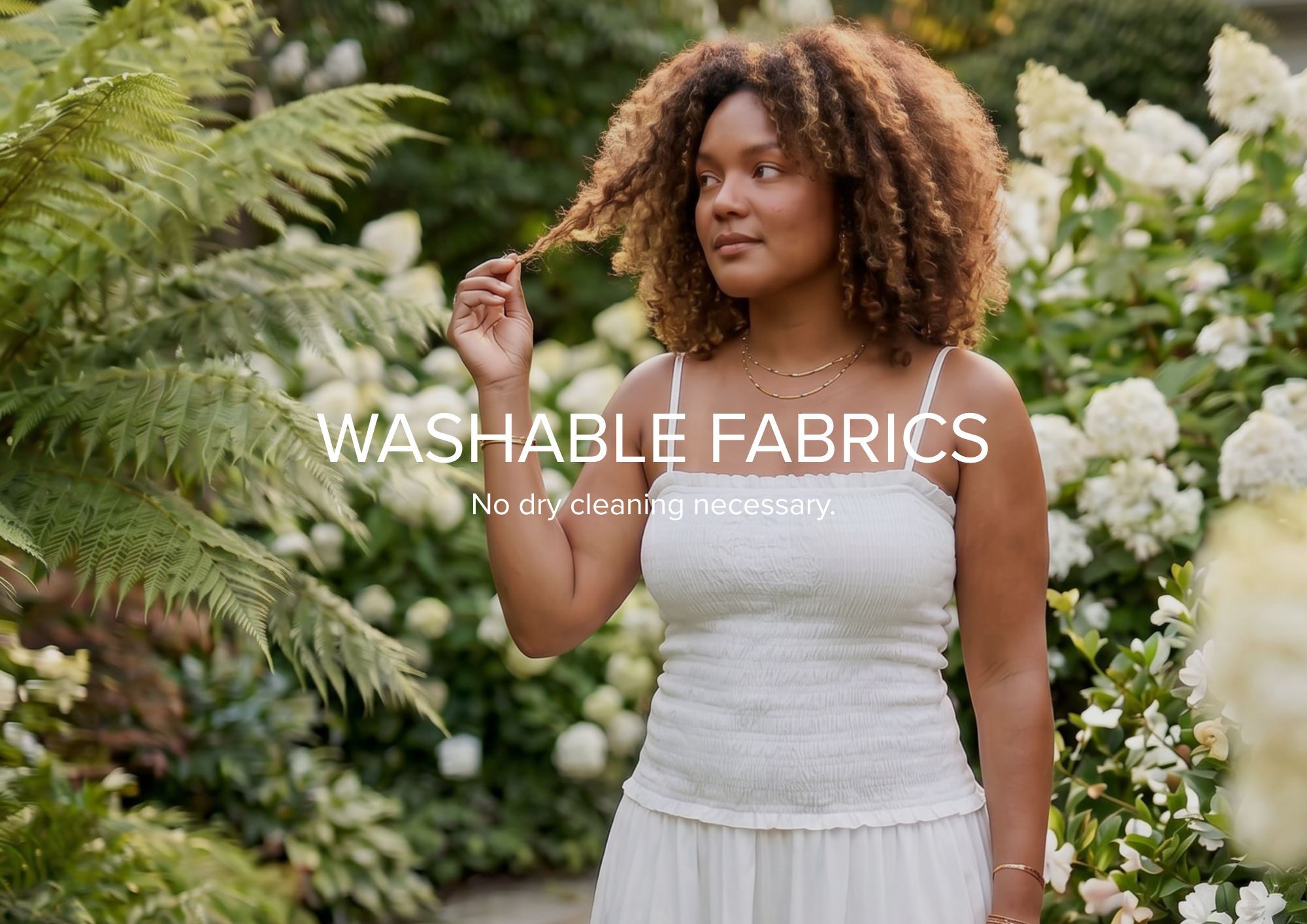 A woman standing outdoors in a garden wearing a white sleeveless top and white skirt, with overlaid text reading 'WASHABLE FABRICS' and 'No dry cleaning necessary.'.