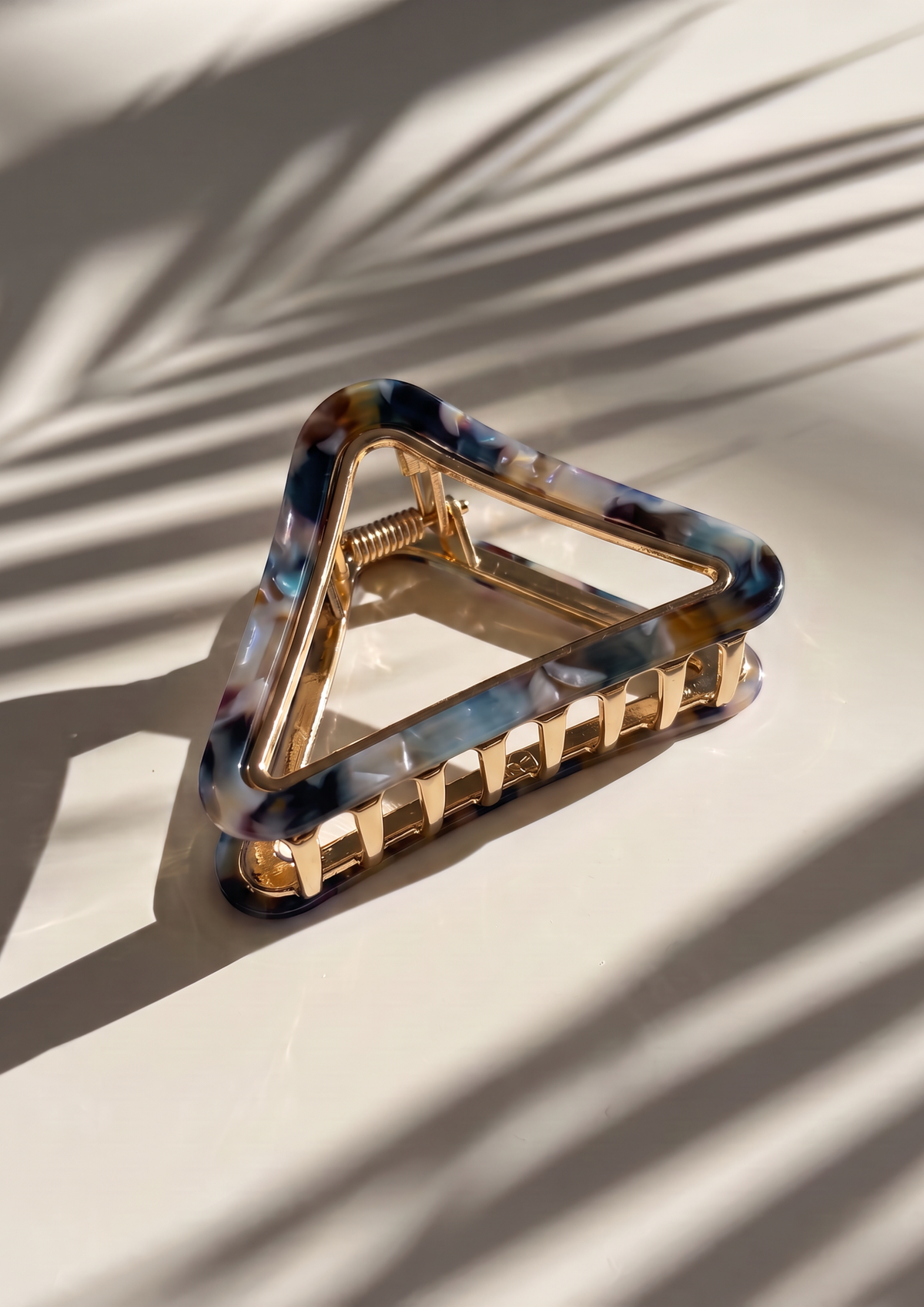 A triangular hair claw clip with a gold-tone metal frame and a marbled tortoiseshell acetate front featuring blue, black, and brown patterns, resting on a light beige surface with palm leaf shadows.