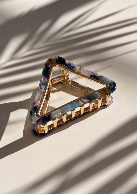 A triangular hair claw clip with a gold-tone metal frame and a marbled tortoiseshell acetate front featuring blue, black, and brown patterns, resting on a light beige surface with palm leaf shadows.