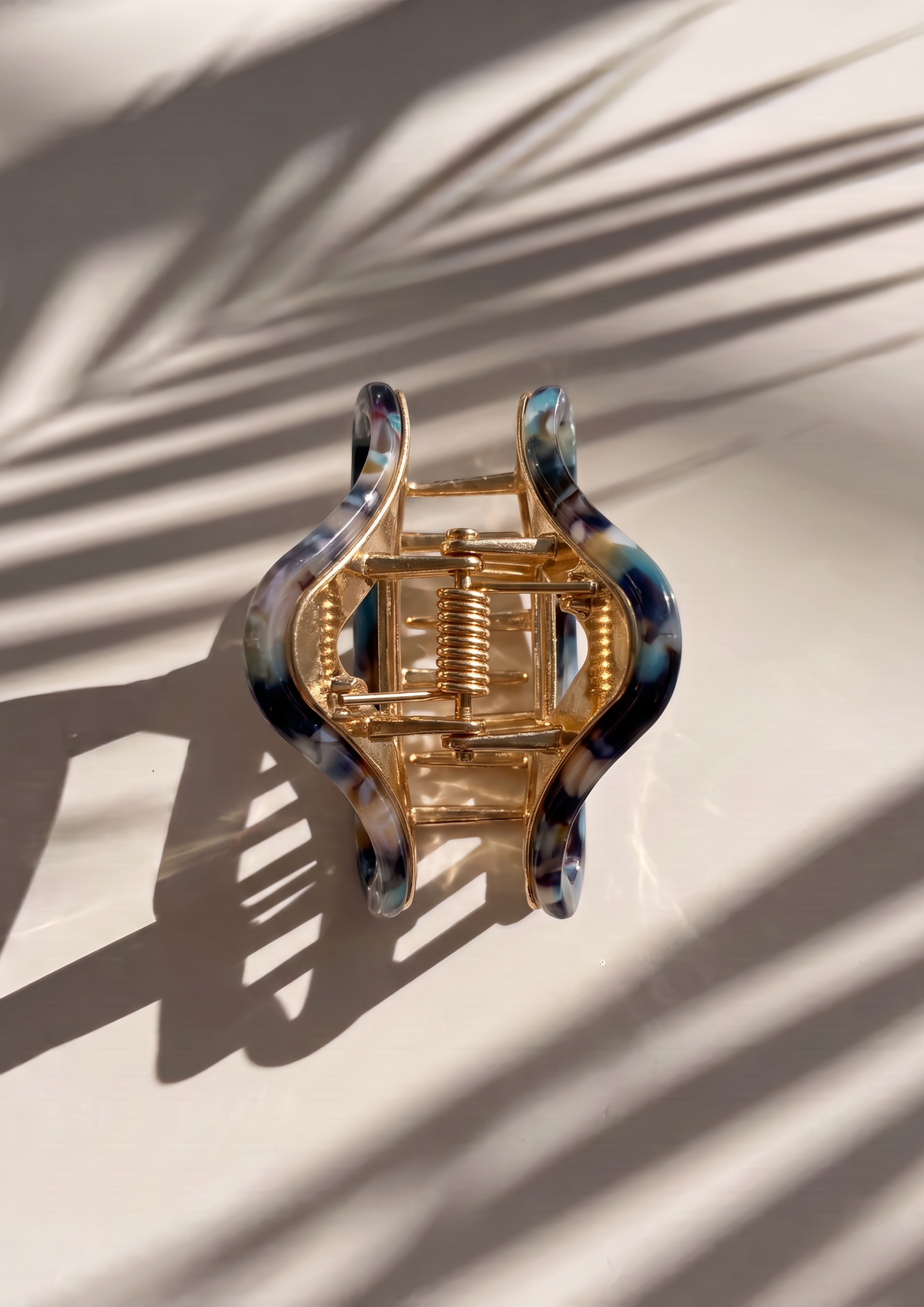 A wavy hair claw clip with a gold-tone metal spring and marbled blue, black, and white resin outer edges, photographed on a light surface with palm leaf shadows.
