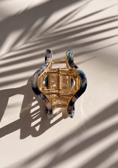 A wavy hair claw clip with a gold-tone metal spring and marbled blue, black, and white resin outer edges, photographed on a light surface with palm leaf shadows.