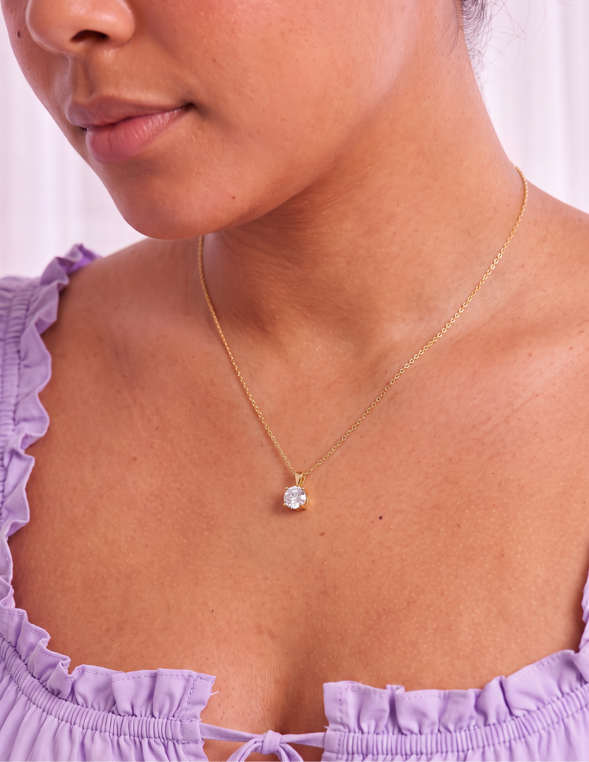 A close-up of a woman wearing a delicate gold chain necklace with a single round clear gemstone pendant, styled with a light purple ruffled top.
