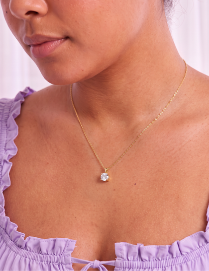A close-up of a woman wearing a delicate gold chain necklace with a single round clear gemstone pendant, styled with a light purple ruffled top.