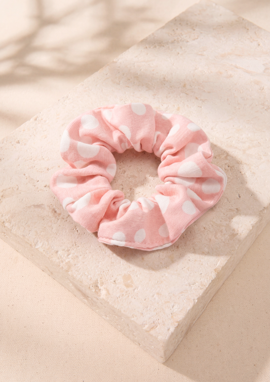 A pink fabric scrunchie with white polka dots, resting on a textured beige stone block with soft shadows.