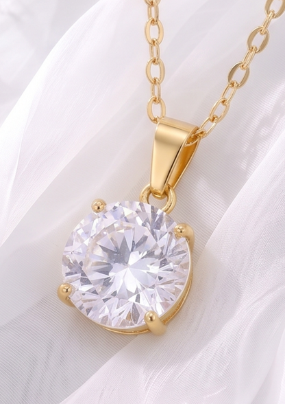 A gold pendant necklace featuring a large, round, clear faceted gemstone in a four-prong setting, resting on a soft white draped fabric background.